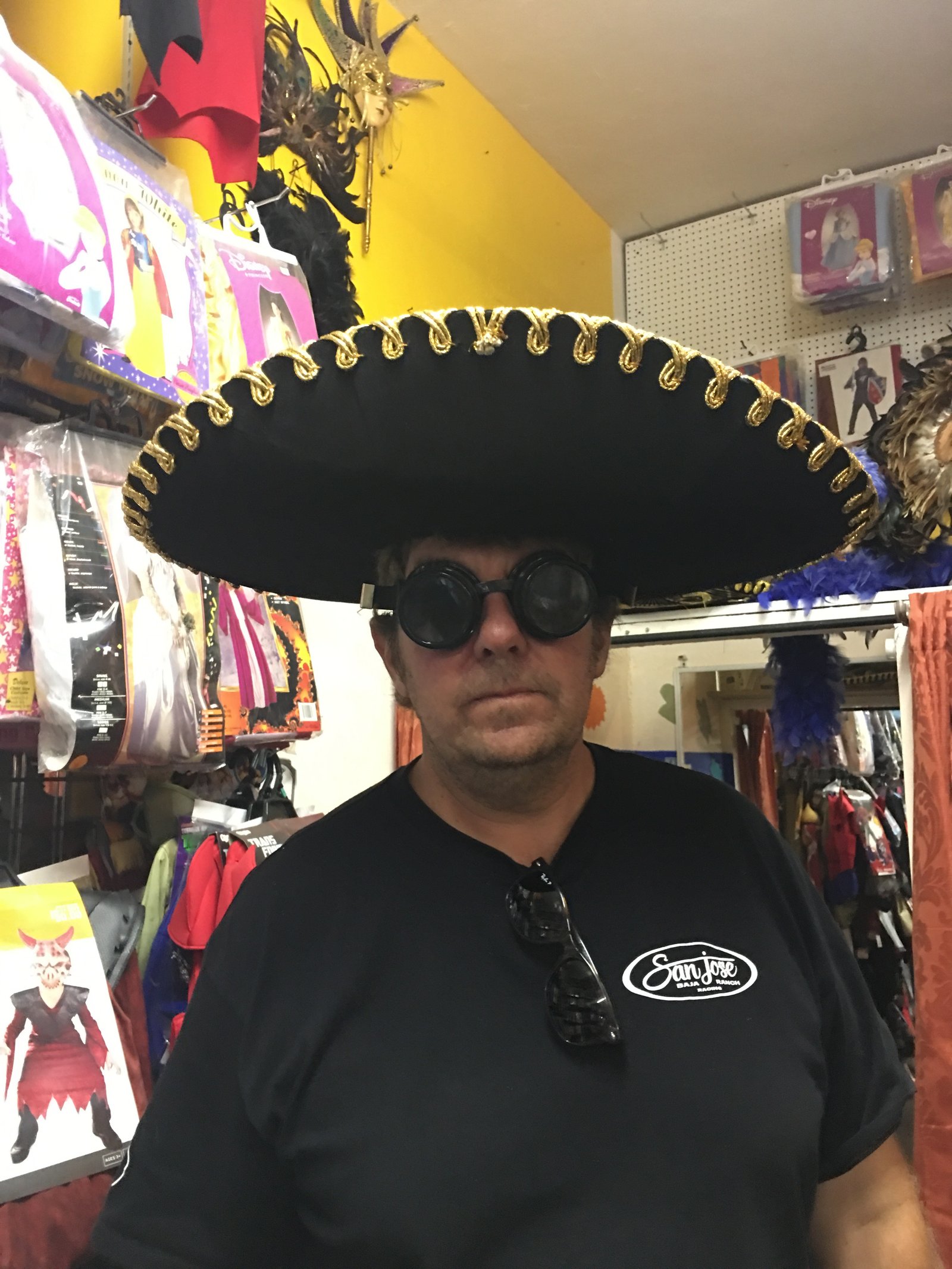 Jay Christopher Humphreys wearing a large black sombrero with gold trim and dark round goggles in a costume shop.