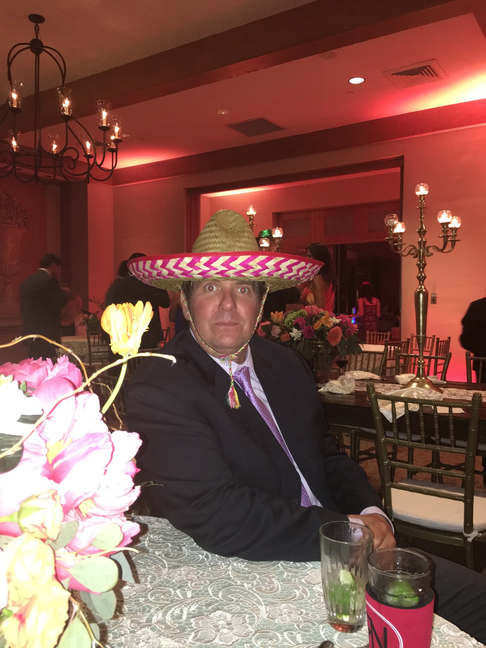 Jay Christopher Humphreys in a suit wearing a woven sombrero with pink and white trim at a formal event table with flowers.