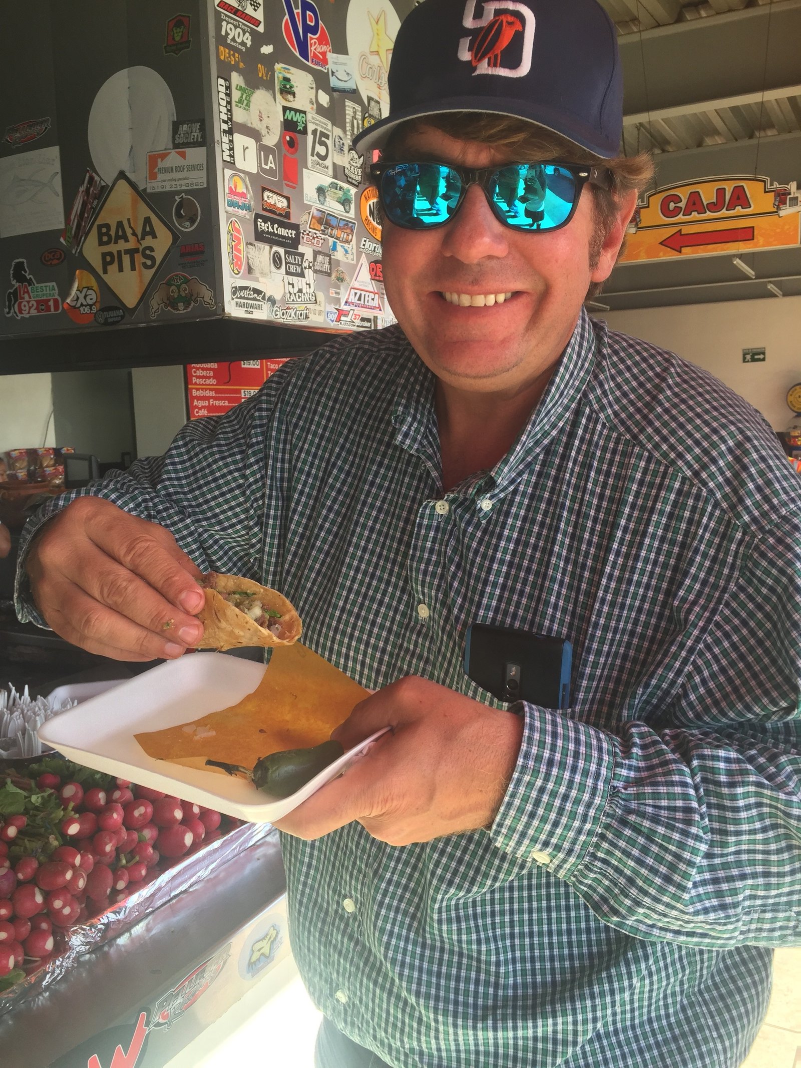 Jay Christopher Humphreys in a checkered shirt and sunglasses holding a taco at a counter restaurant.